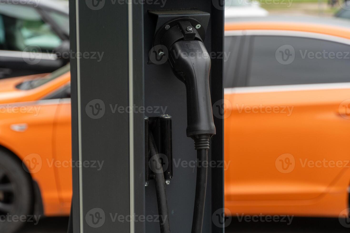 Close view of an electric vehicle charging station showcasing a connector in use, with a bright orange car parked nearby in a modern urban setting photo