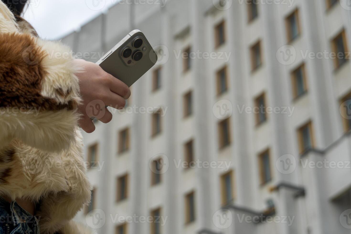 A person dressed in a stylish faux fur coat is focused on their smartphone in a contemporary urban setting featuring an impressive building with large windows photo