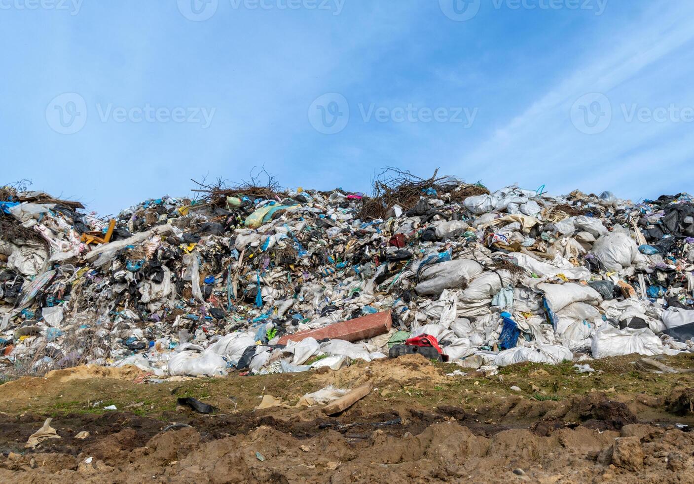 A large mound of garbage is visible at a landfill, consisting of various waste types including plastic bags and discarded furniture, illuminated by sunlight on a clear day photo