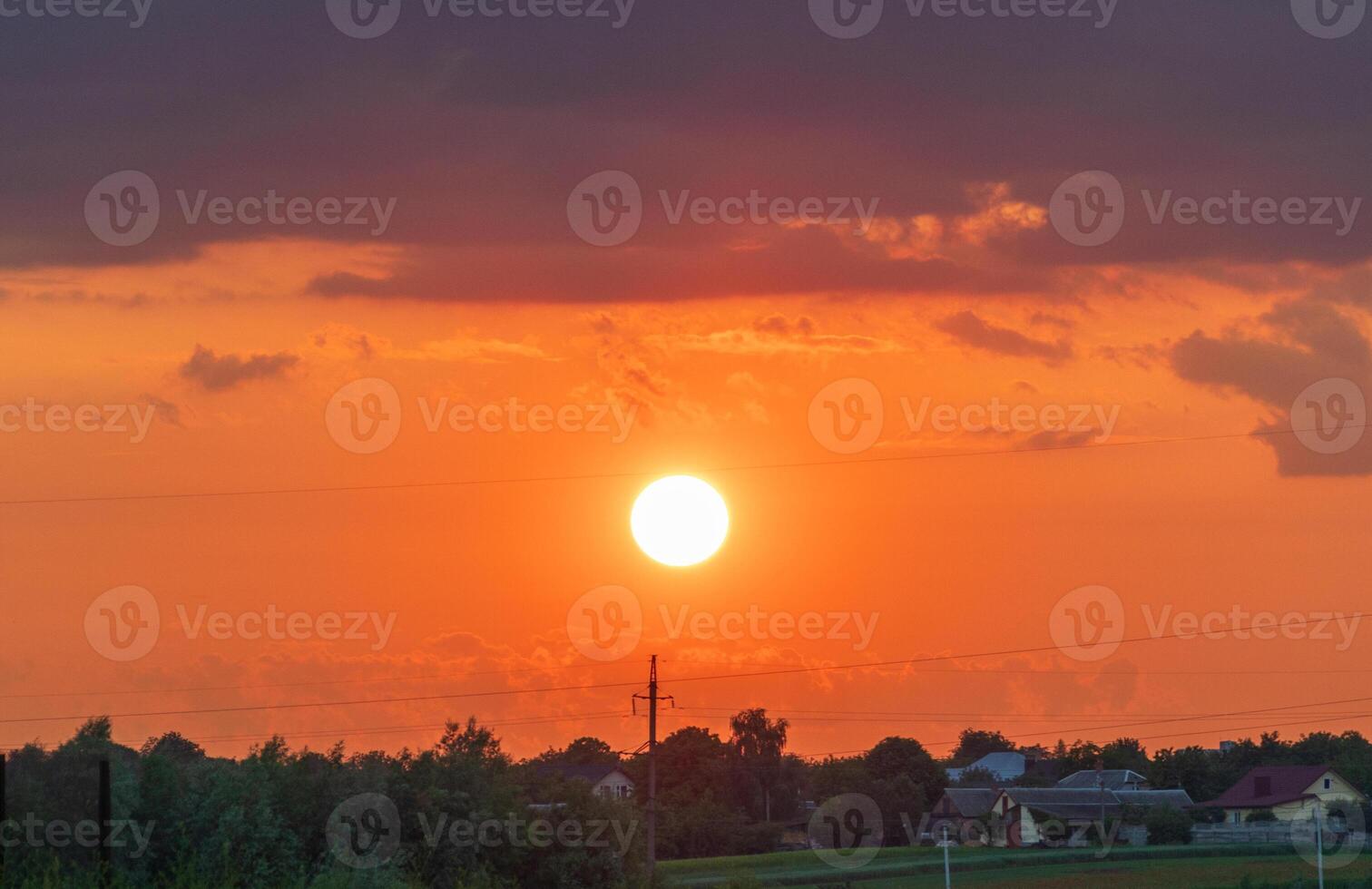 Vibrant sunset casts an orange glow over a rural landscape. Silhouettes of houses and trees create a peaceful atmosphere as day transitions into night photo