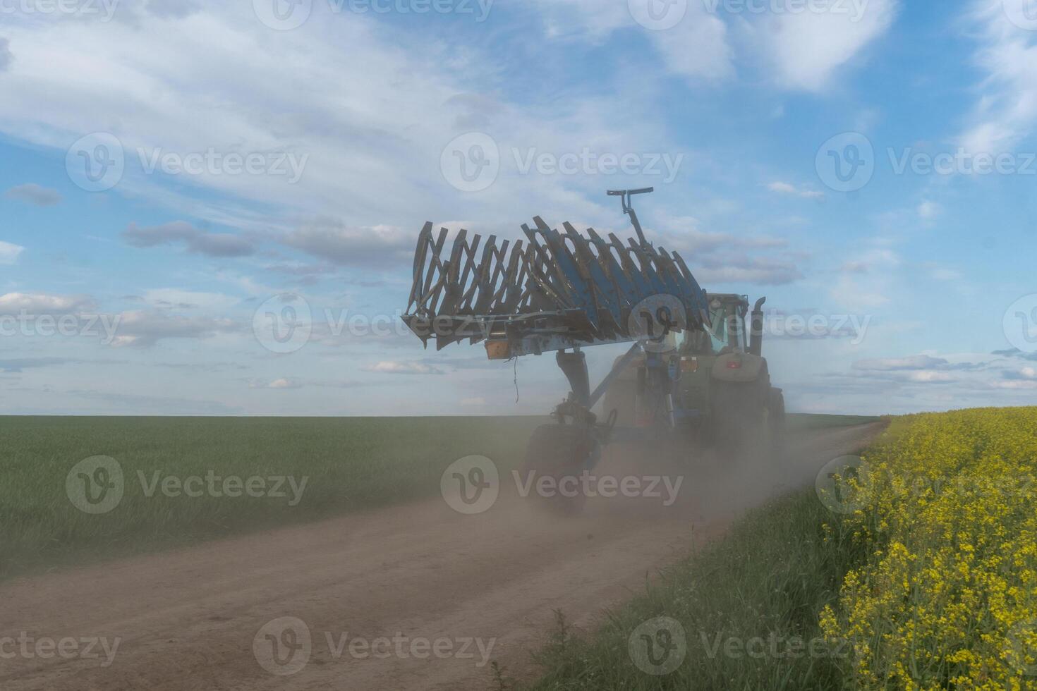 A tractor is plowing a dusty dirt road in a vast field, with patches of yellow flowers and a vibrant sky in the background during the golden hour of sunset photo