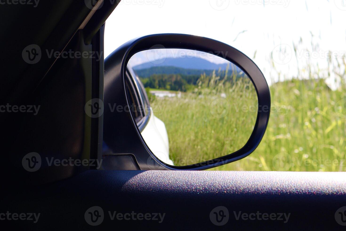A side mirror reflects a green field and forested hills under a bright sky, captured from inside a car to convey a peaceful travel moment in nature. photo