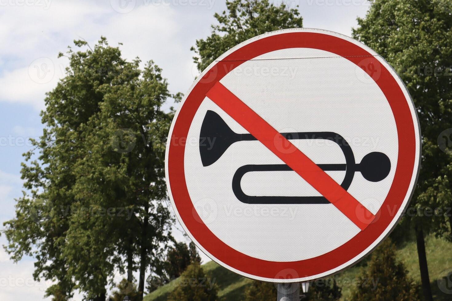 Close-up of a no honking traffic sign with a horn crossed out in red. The round sign stands against a background of green trees and a partly cloudy sky. photo