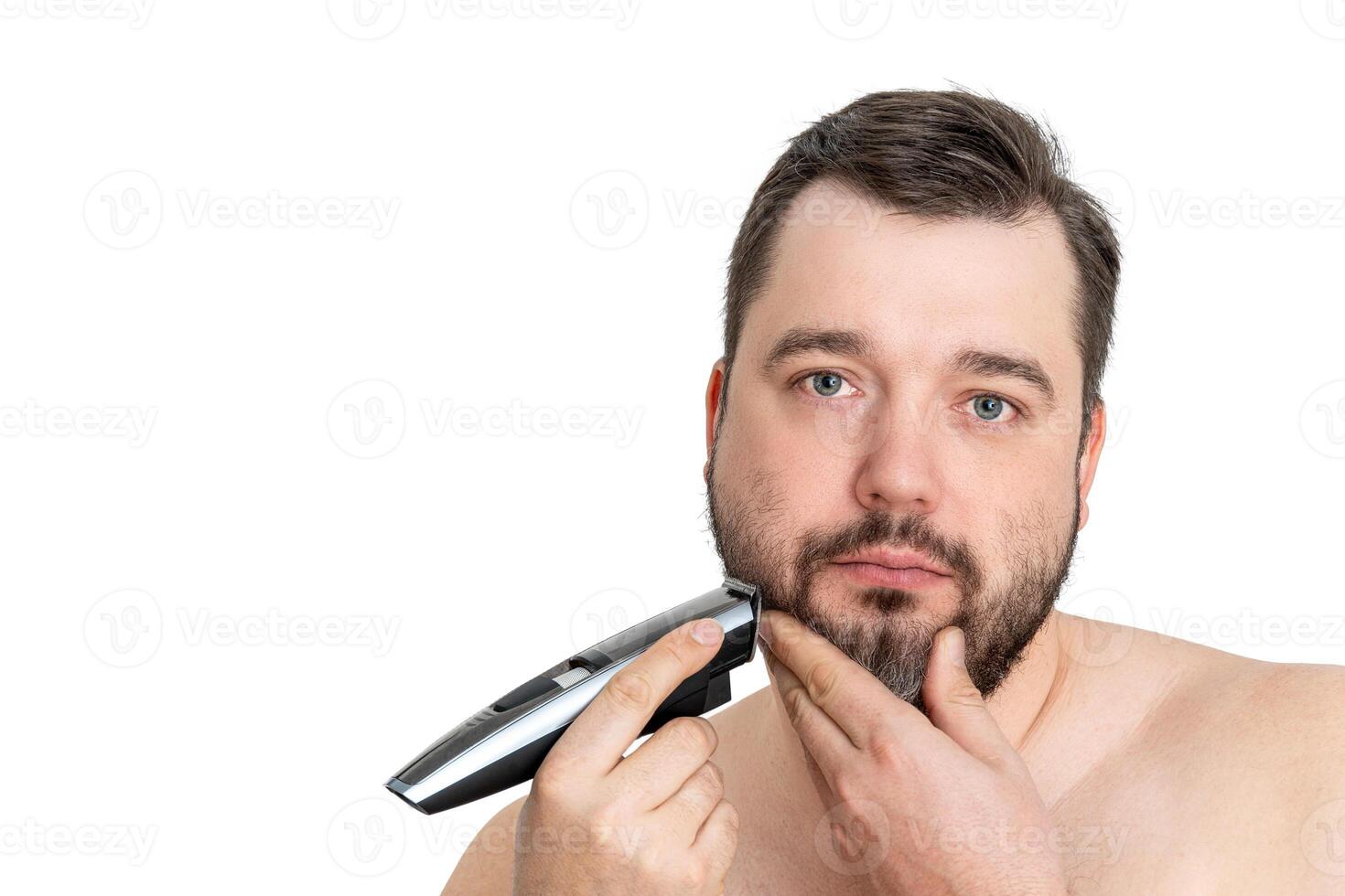A young man with casual clothing is using an electric shaver to groom and shave his well-maintained beard. The image showcases personal grooming and hygiene against a clean white background. photo