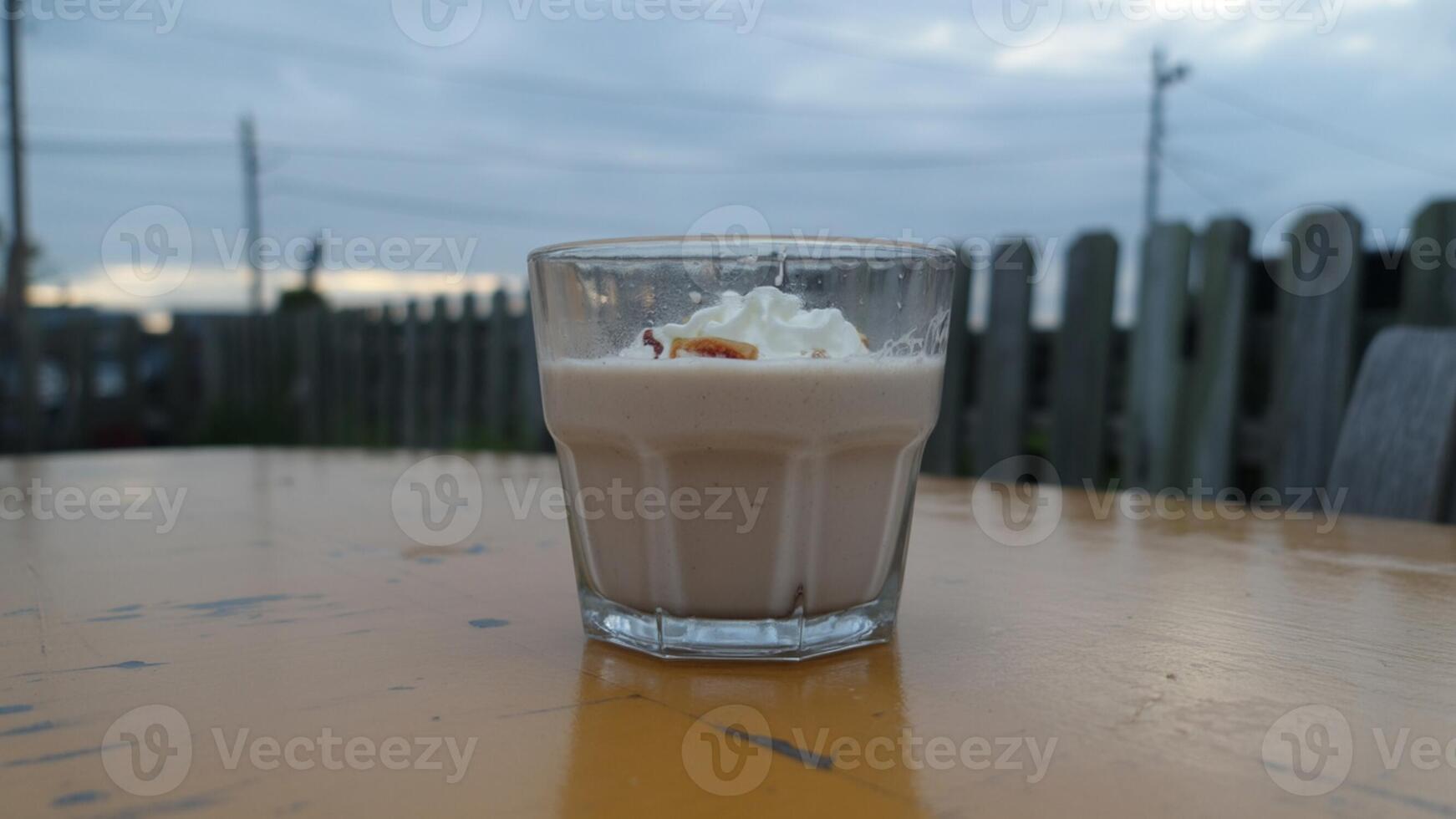 A glass of dessert with whipped cream and a topping on a yellow table with a cloudy sky behind it photo