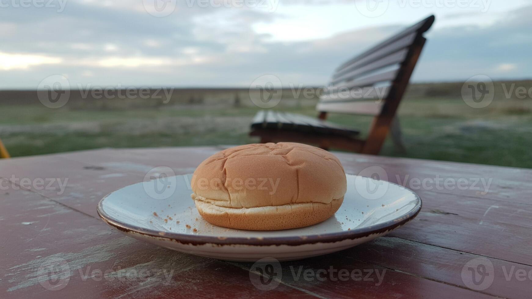 A burger bun on a plate sitting on a picnic table with a bench and a field in the background photo