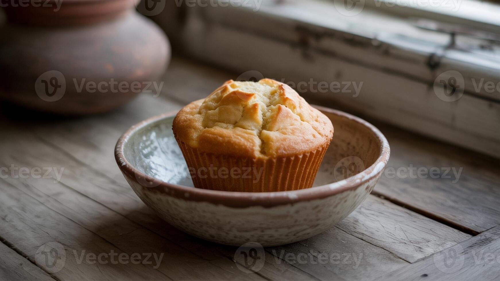 A single muffin in a bowl on a wooden surface near a window with a clay pot in the background photo