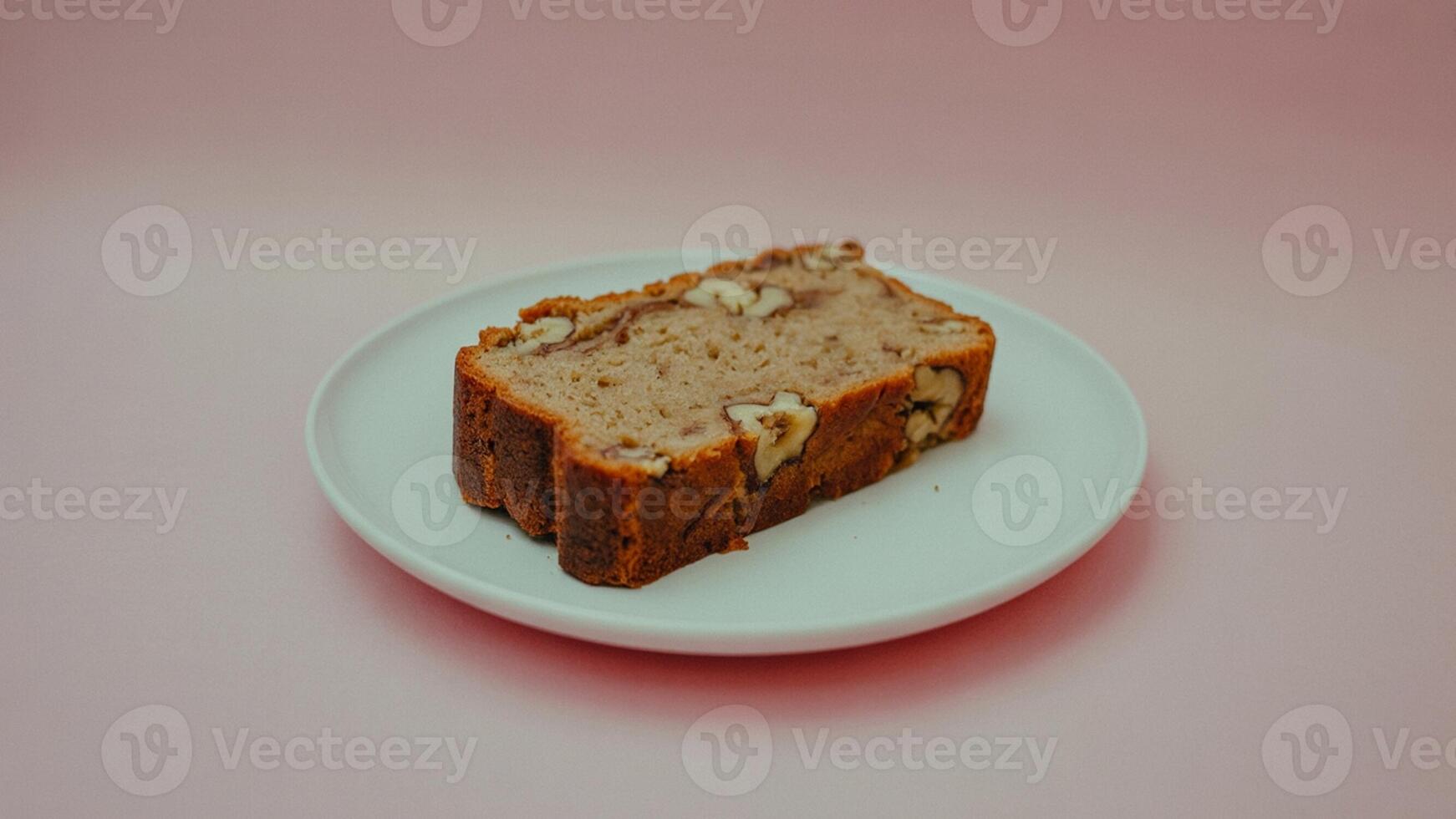 A slice of banana nut bread on a white plate against a light pink background in a studio setting photo