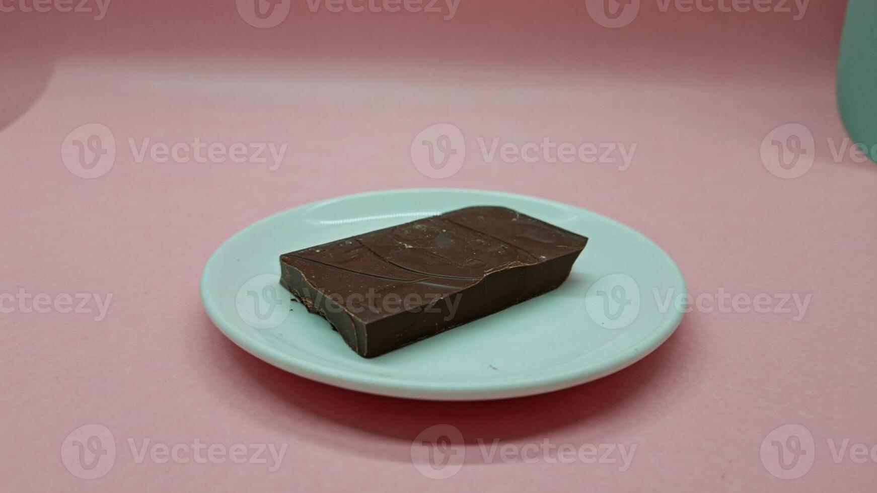 A rectangular piece of chocolate on a small white plate against a pink background in a studio shot photo