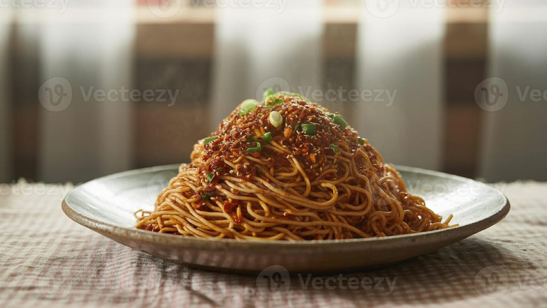 A large plate of noodles topped with sauce and green onions on a patterned surface near a window photo