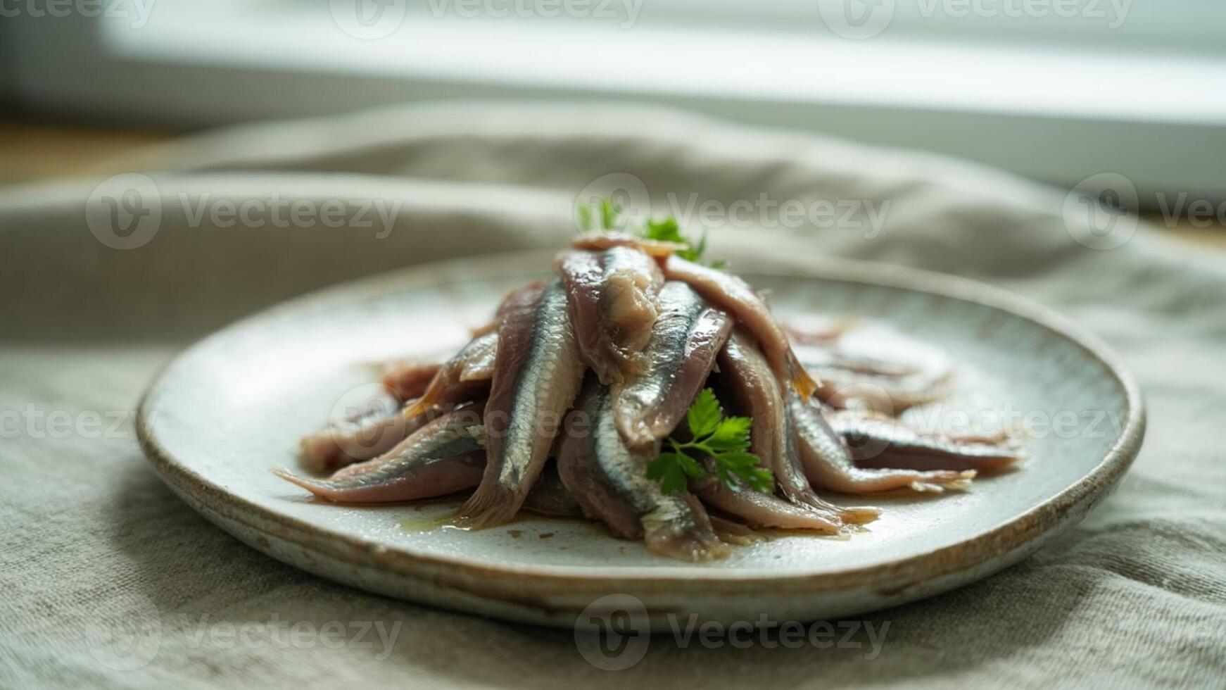 A pile of anchovies on a plate garnished with parsley sitting on a linen cloth with a window behind photo