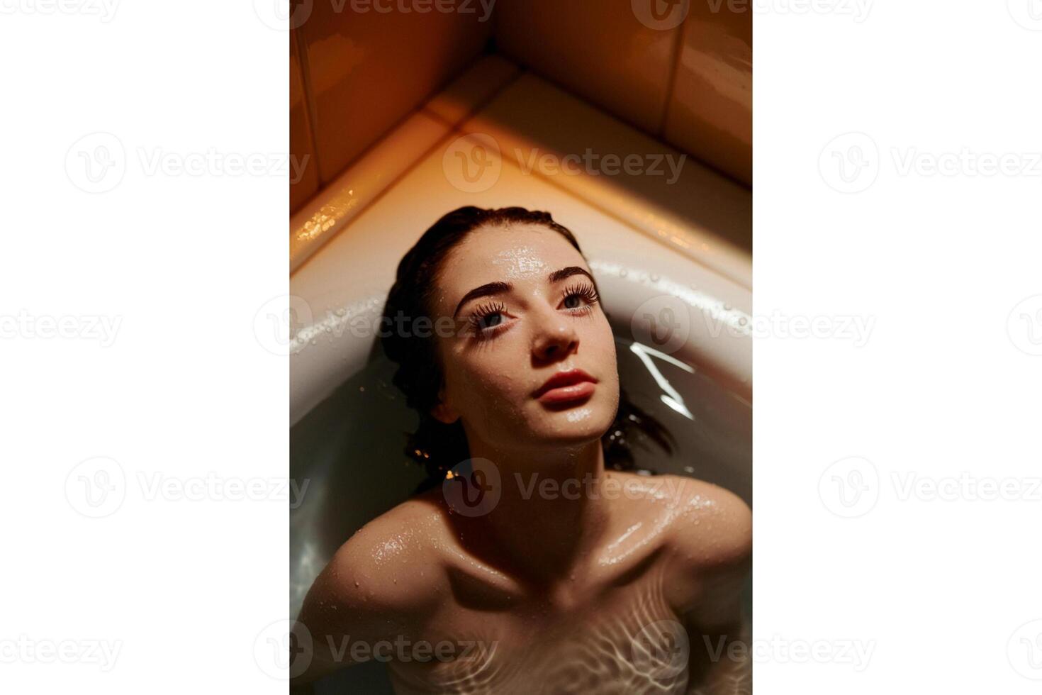 A woman with dark hair lying in a bathtub looking up with water on her skin photo