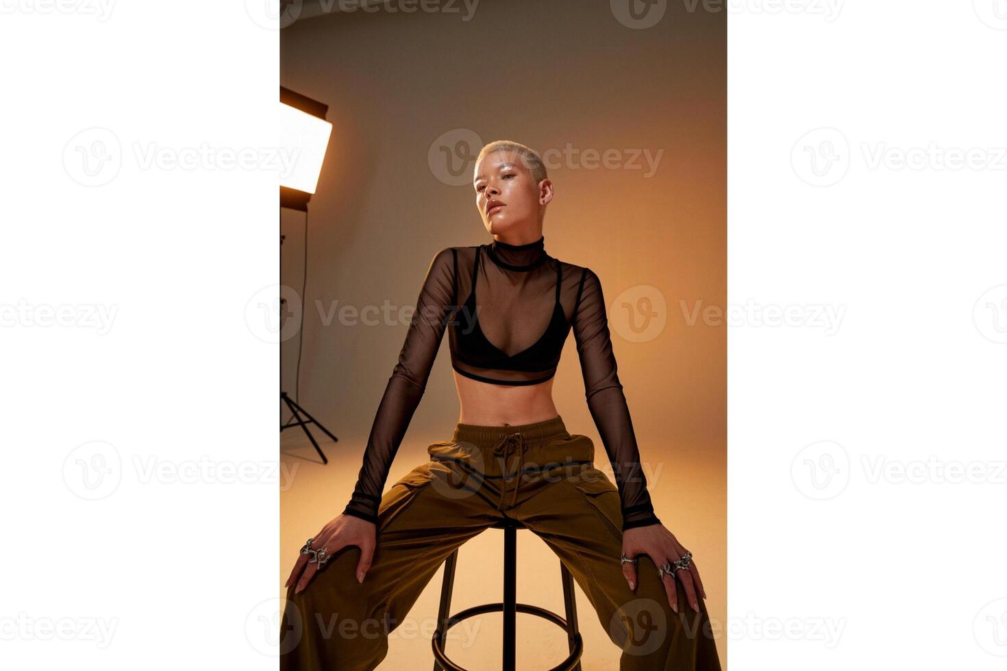 Woman with shaved head sitting on a stool wearing a mesh top and cargo pants photo