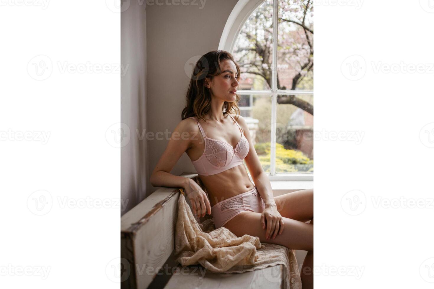 Woman in pink lingerie sitting near window with tree branches outside looking away photo