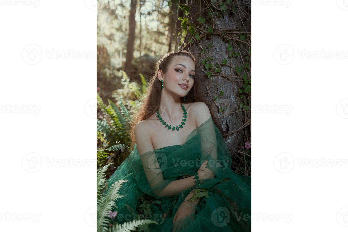 Portrait of a woman in green dress and jewelry sitting by a tree in a forest photo