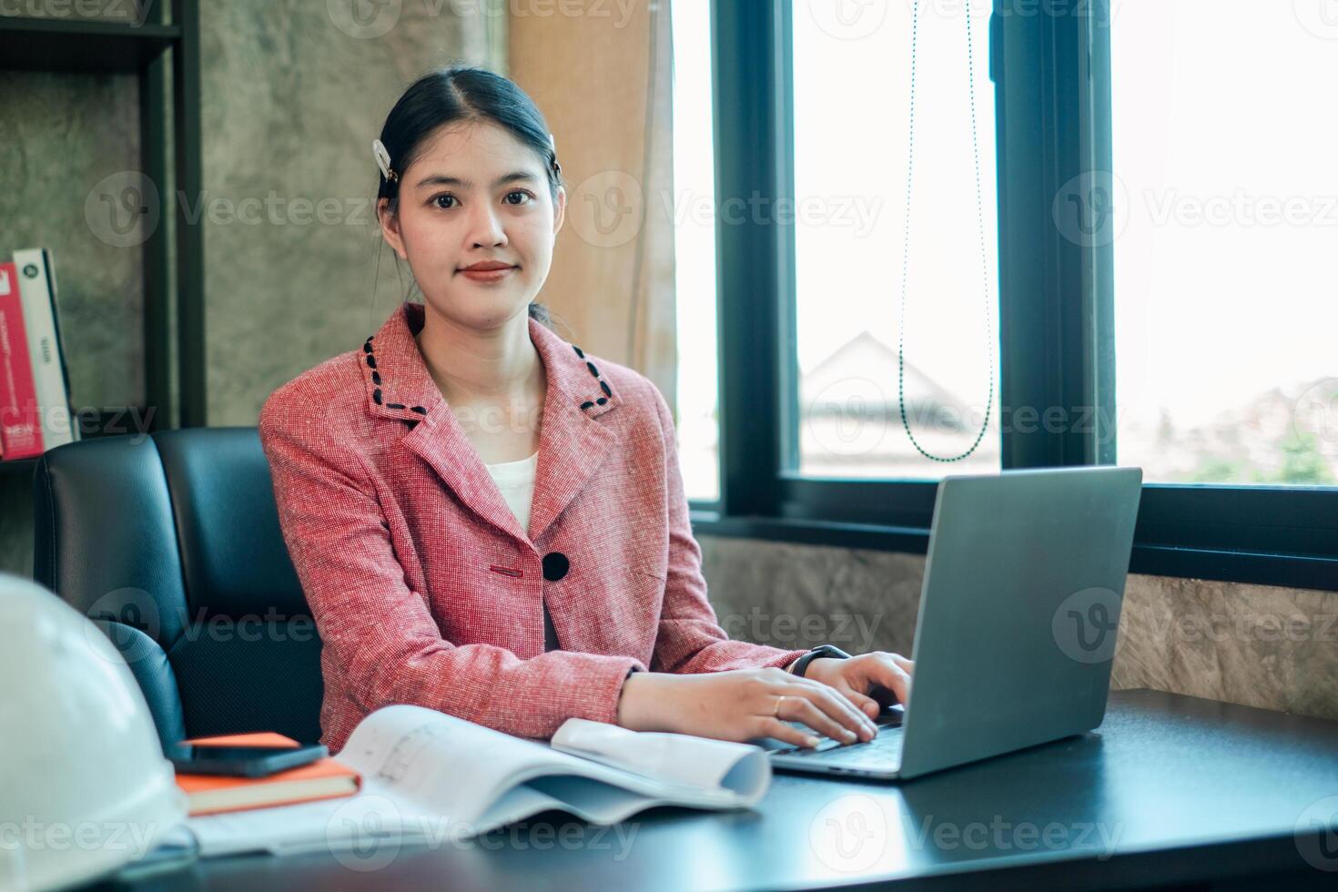 Businesswoman in pink blazer using laptop at office desk, surrounded by documents and a hard hat, near a large window. photo