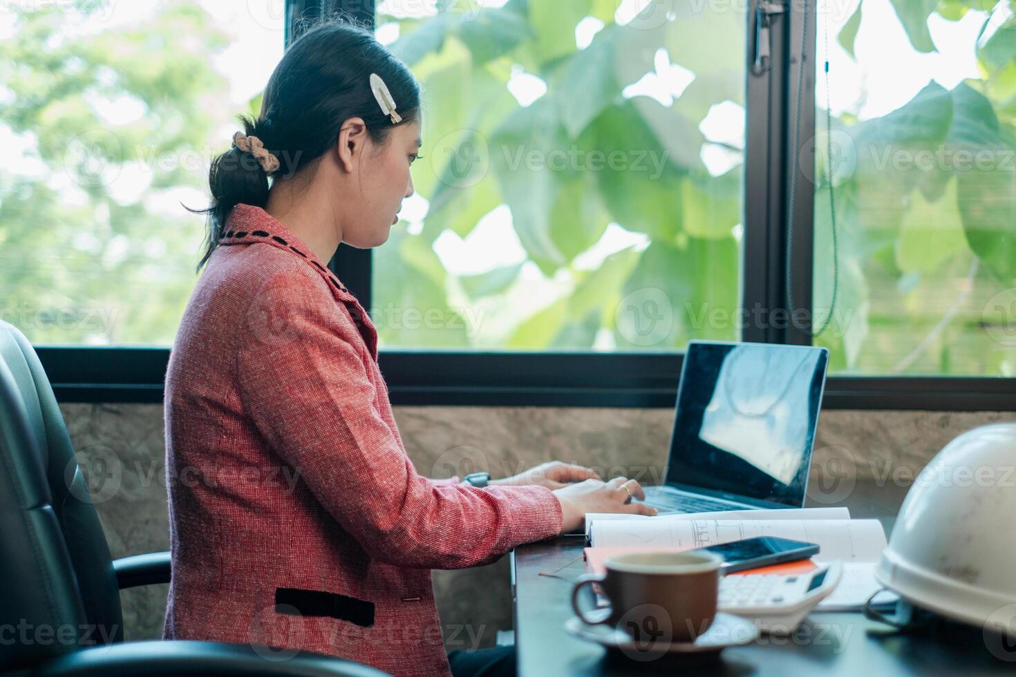 Businesswoman focused on laptop in a bright office, surrounded by documents and coffee, with a view of lush greenery. photo