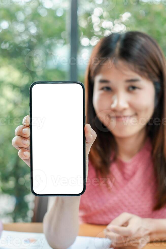 Smiling woman presenting a smartphone with a blank screen, ideal for app or website mockup, in a bright room with greenery outside. photo
