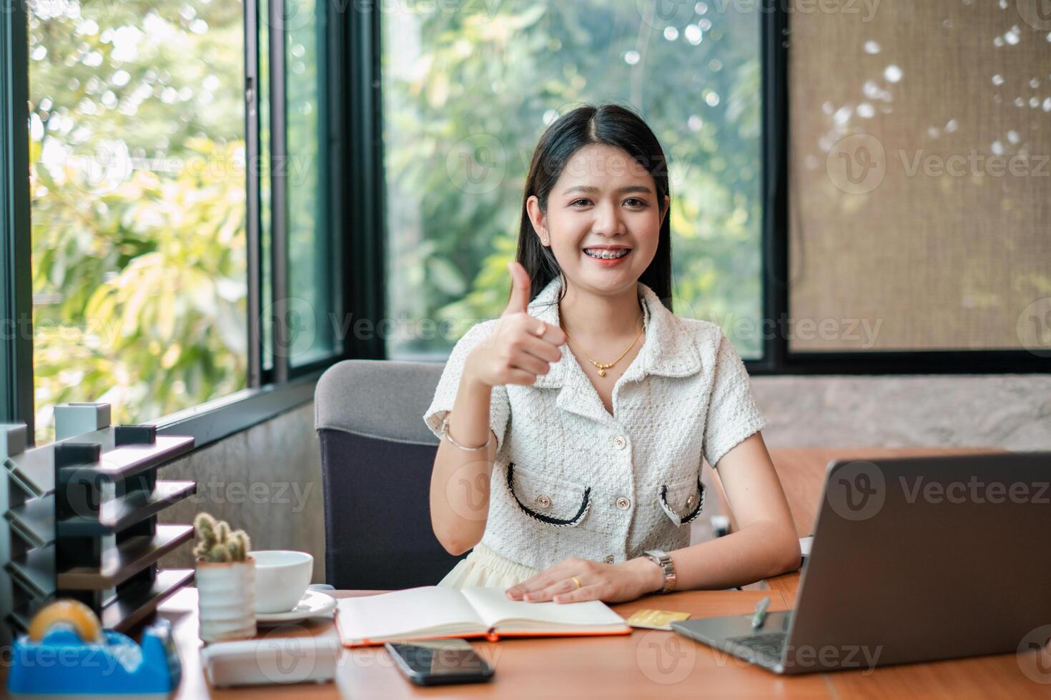 Smiling young woman in office giving thumbs up, sitting at desk with laptop and documents, showcasing positivity and success. photo