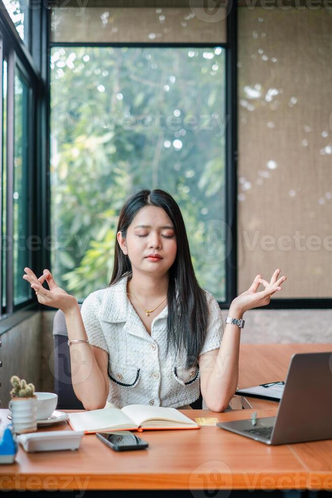 A young woman practices meditation at her office desk, surrounded by a laptop and notebook, in a bright and modern workspace. photo