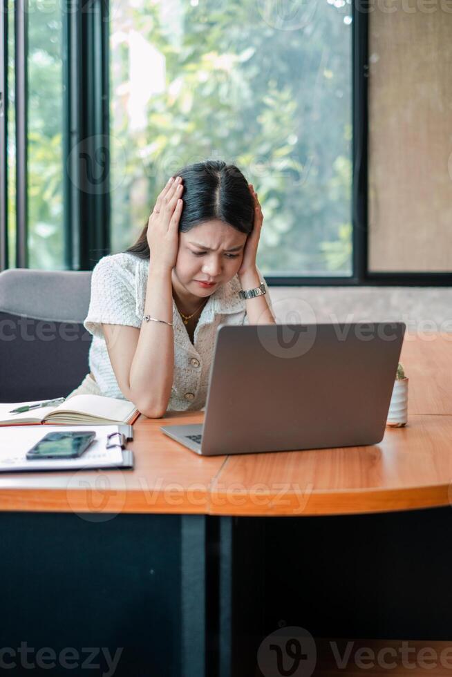 A businesswoman looks stressed while working on her laptop at a modern office desk, surrounded by documents and a smartphone. photo