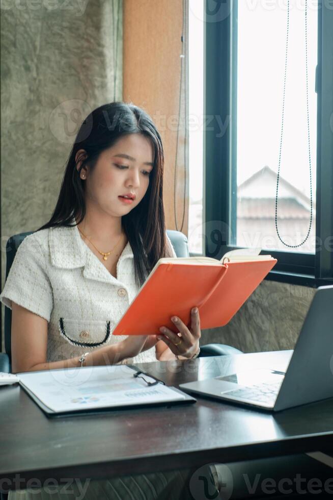 A young woman sits at a desk in a modern office, reading a book with a laptop nearby, illuminated by natural light from a window. photo