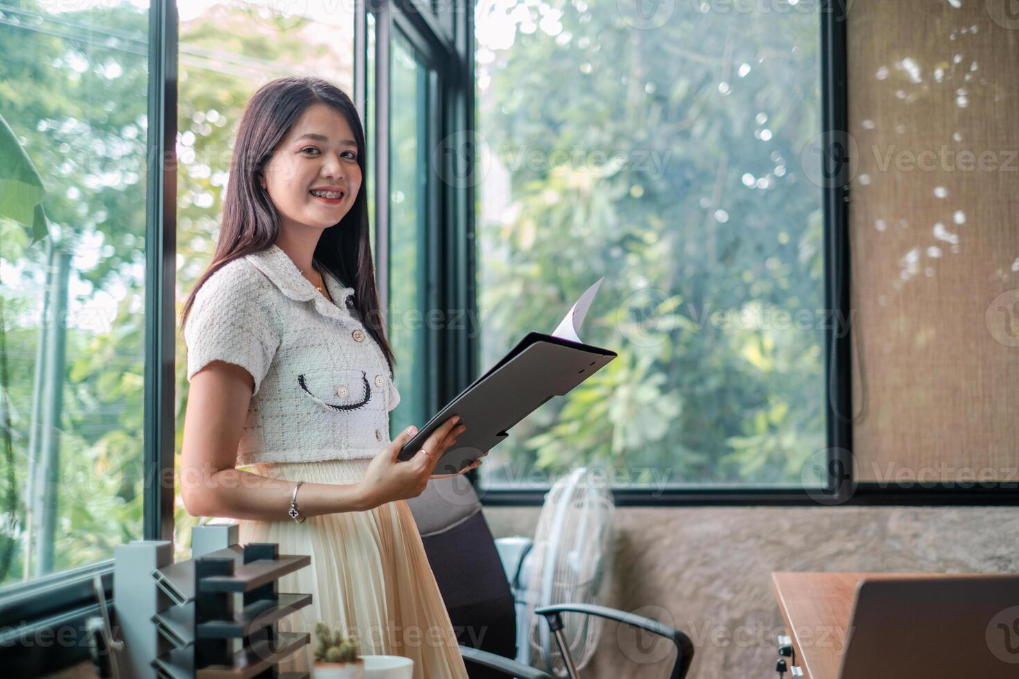 Smiling young woman in a modern office, holding documents, standing by large windows with natural light. photo