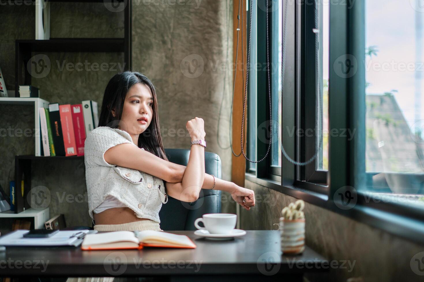 Young woman stretches at her desk, enjoying a break with coffee and an open notebook in a modern office setting. photo