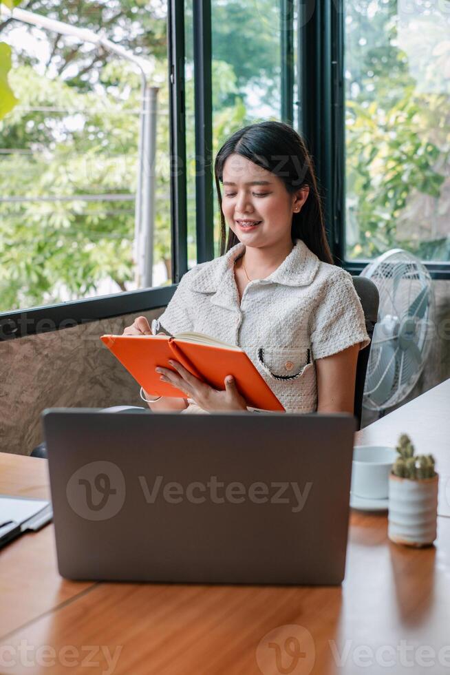 A young woman working on a laptop and taking notes in a bright, modern office with large windows and greenery outside. photo