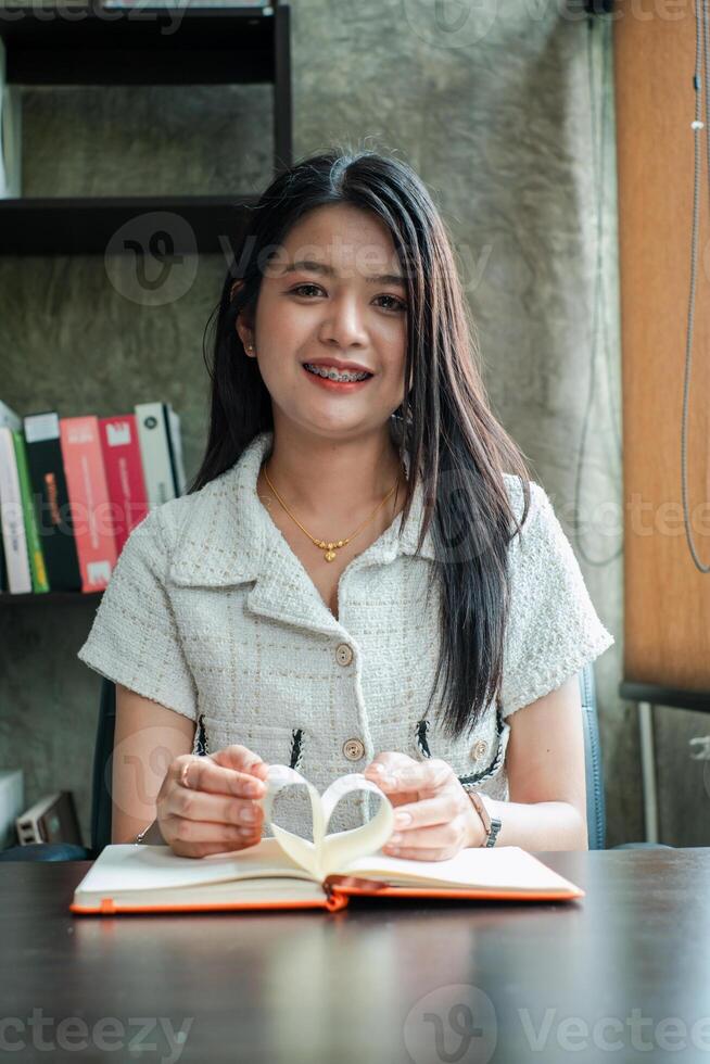 A young woman with long hair smiles while reading a book in a cozy office environment, featuring a bookshelf in the background. photo