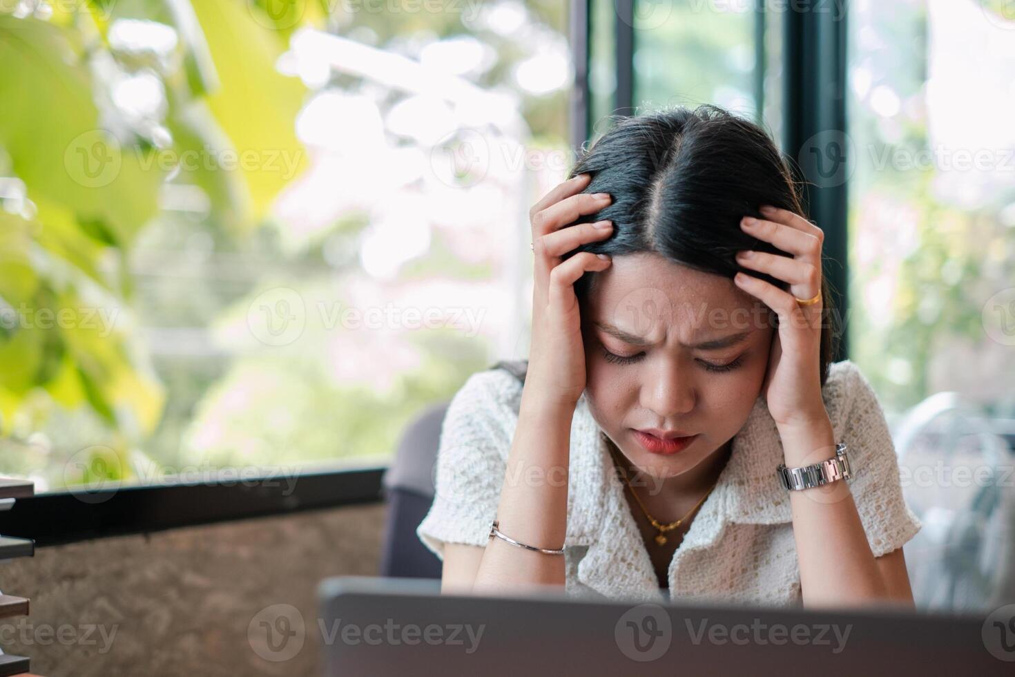 A woman appears stressed while working on a laptop in a modern office setting with natural light and greenery outside. photo