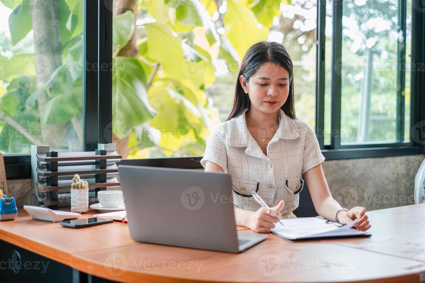A young woman focuses on work at a modern office desk, using a laptop and taking notes near large windows with natural light. photo