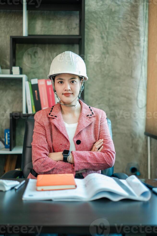 Professional woman engineer wearing a hard hat, sitting at a desk with documents, showcasing confidence and expertise in a modern office setting. photo