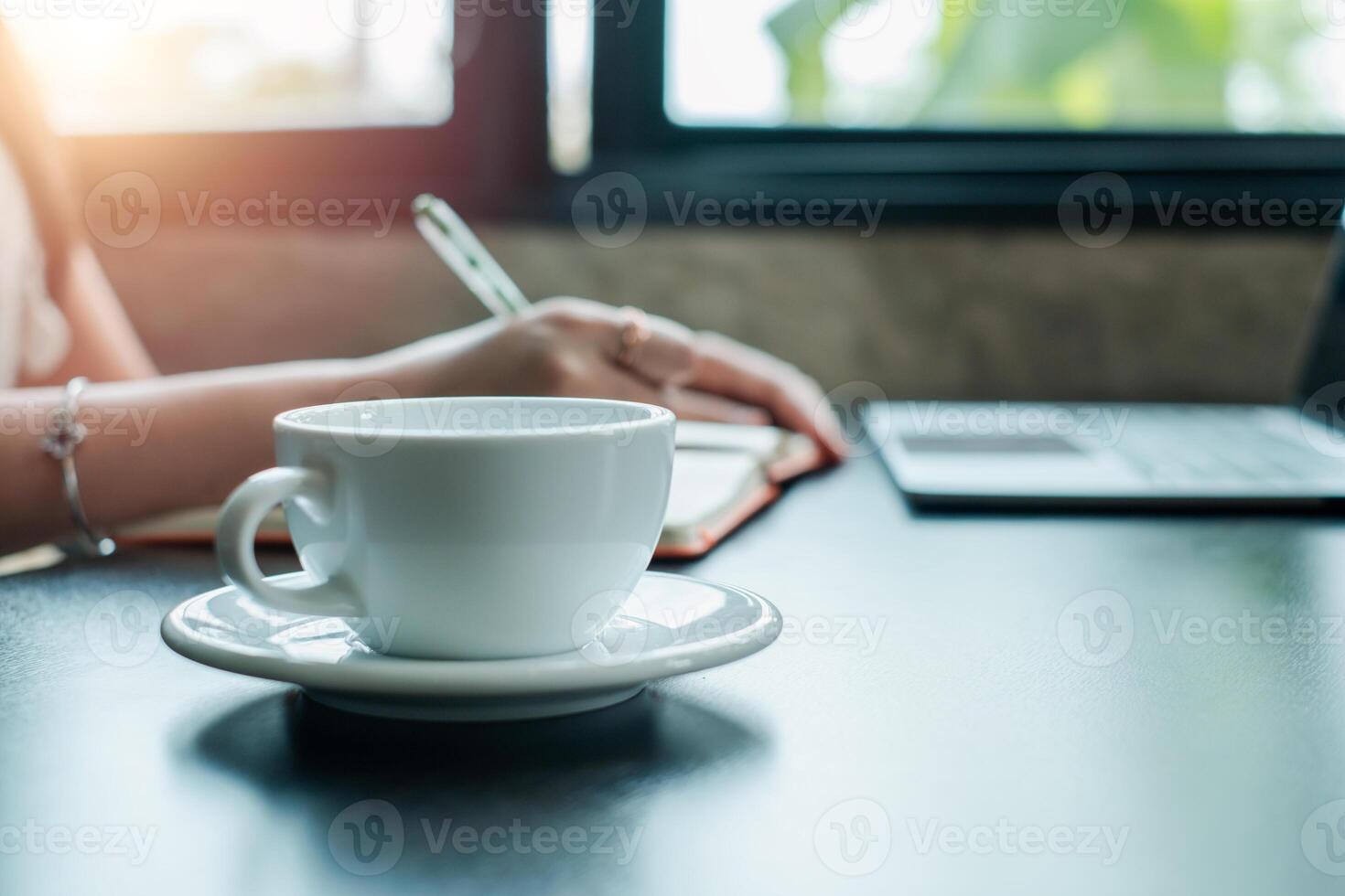A serene morning scene featuring a cup of coffee, a woman writing in a notebook, and a laptop on a desk by the window. photo