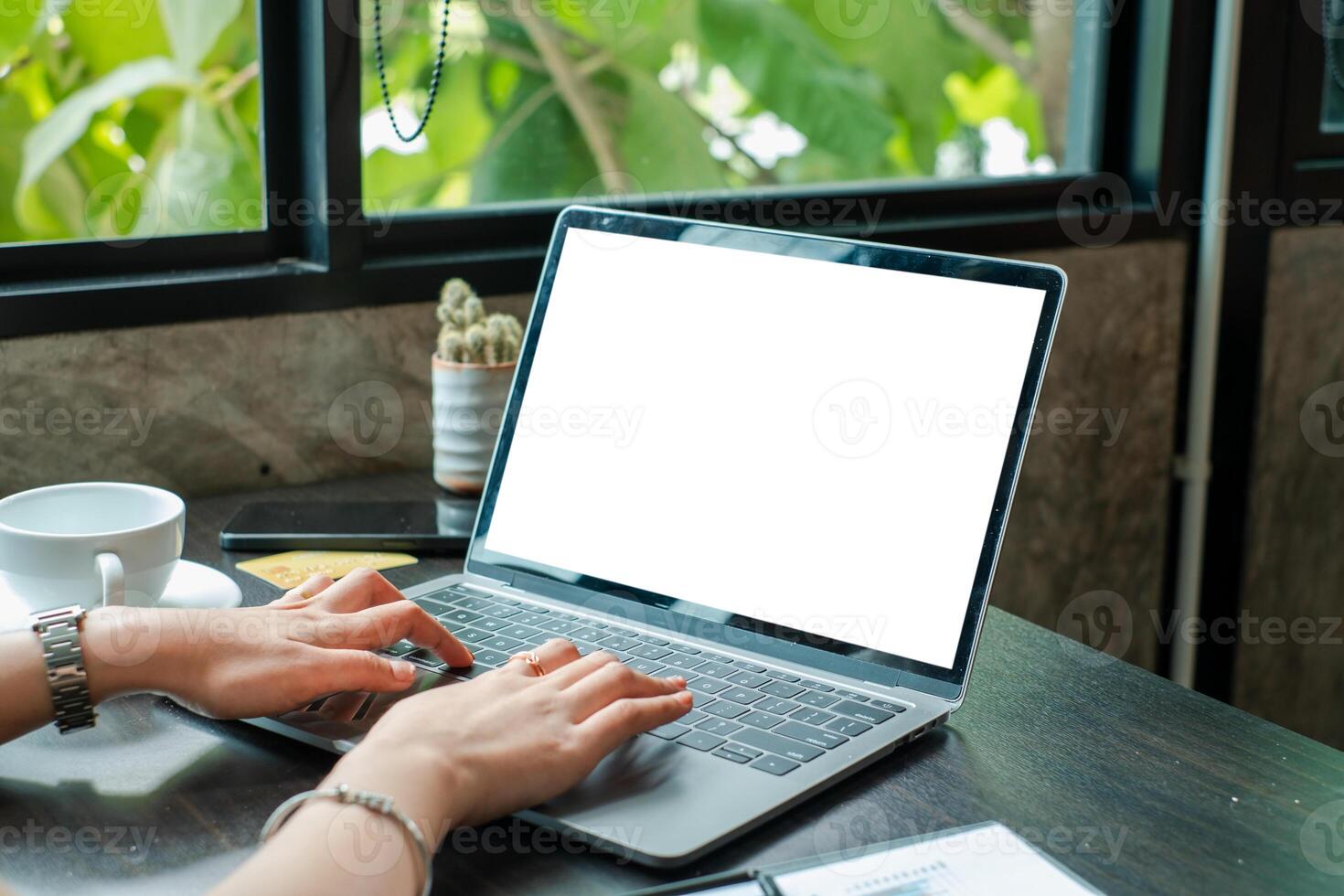 Hands typing on a laptop with a blank screen in a cozy cafe, surrounded by plants and natural light. photo