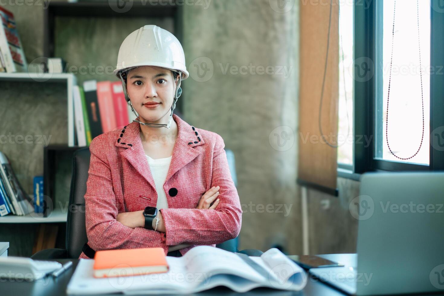 Female engineer in a pink blazer and hard hat, confidently seated at a desk in a modern office setting. photo