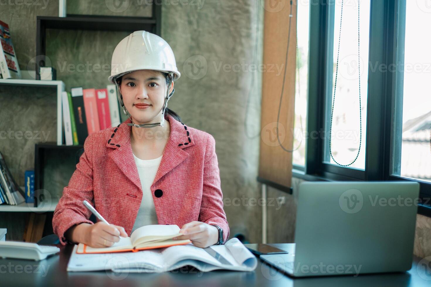 Female engineer wearing a hard hat, working at a desk with a laptop and documents in a modern office setting. photo