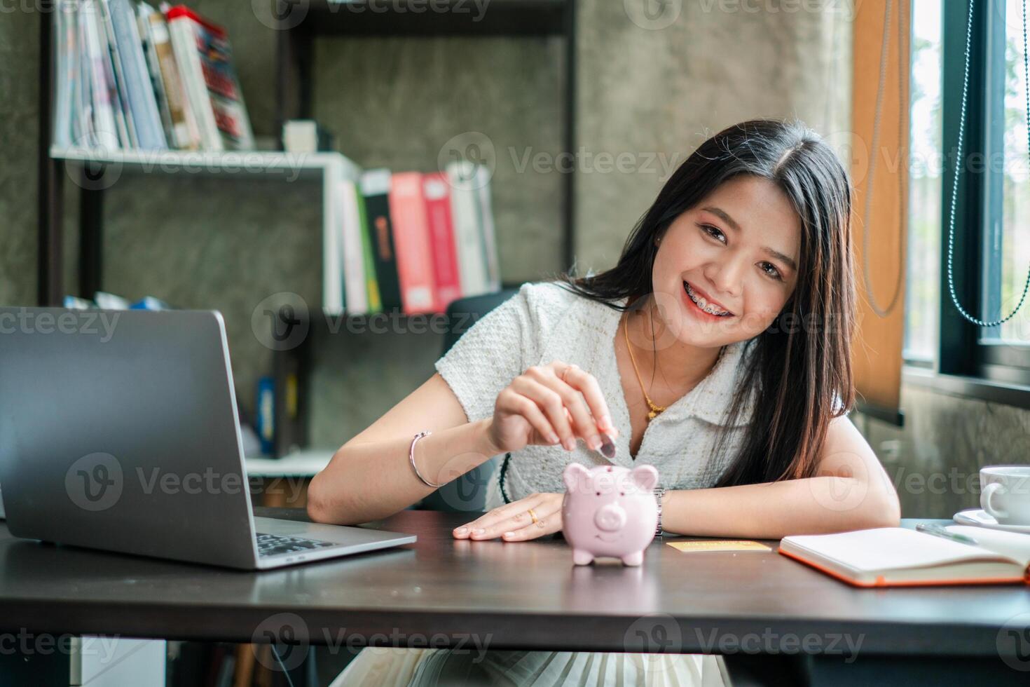 Smiling woman placing coin in piggy bank at desk, symbolizing savings and financial planning. photo