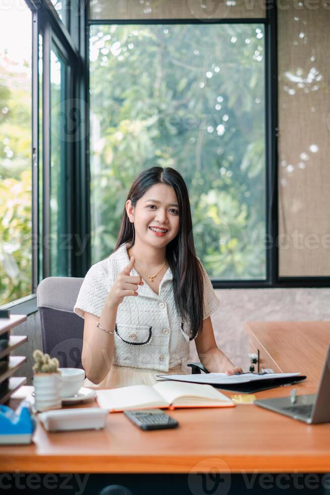 Confident young woman sitting at a desk in a bright office, surrounded by greenery, working on a laptop. photo