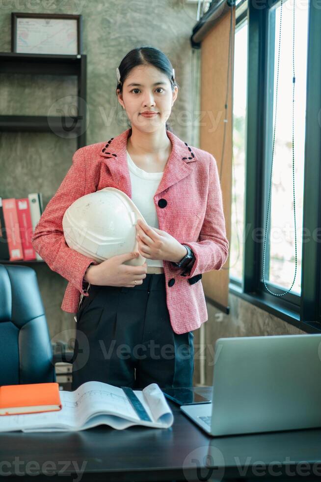 Professional woman engineer in office, holding helmet, with plans and laptop, showcasing modern workplace and career in engineering. photo