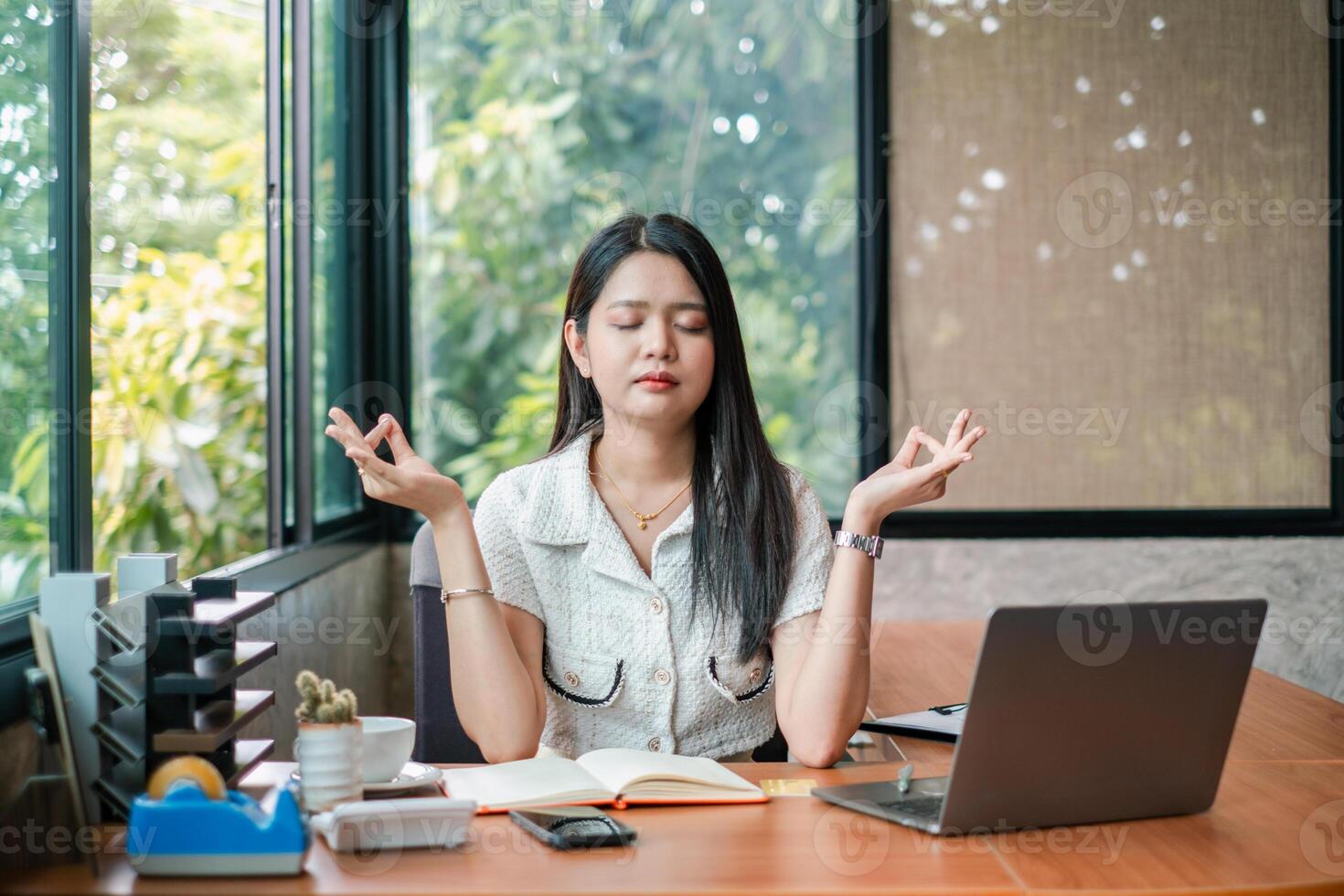 A young woman practices meditation at her desk in a modern office, surrounded by a laptop, notebook, and greenery outside the window. photo