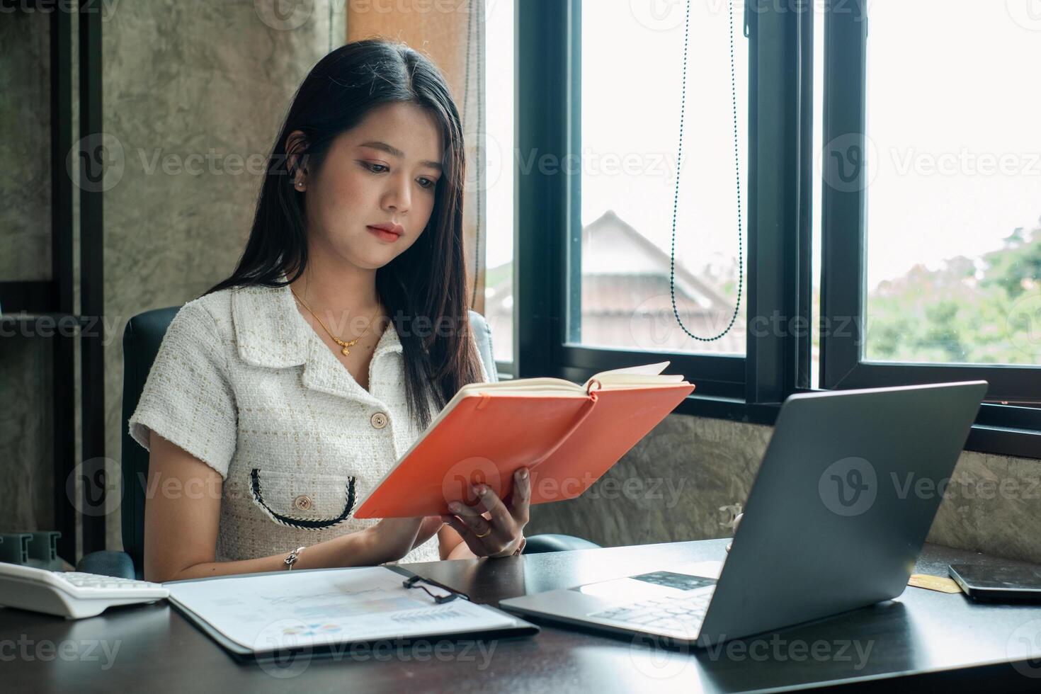 A young woman in a modern office reads a book by the window, with a laptop and documents on the desk. photo