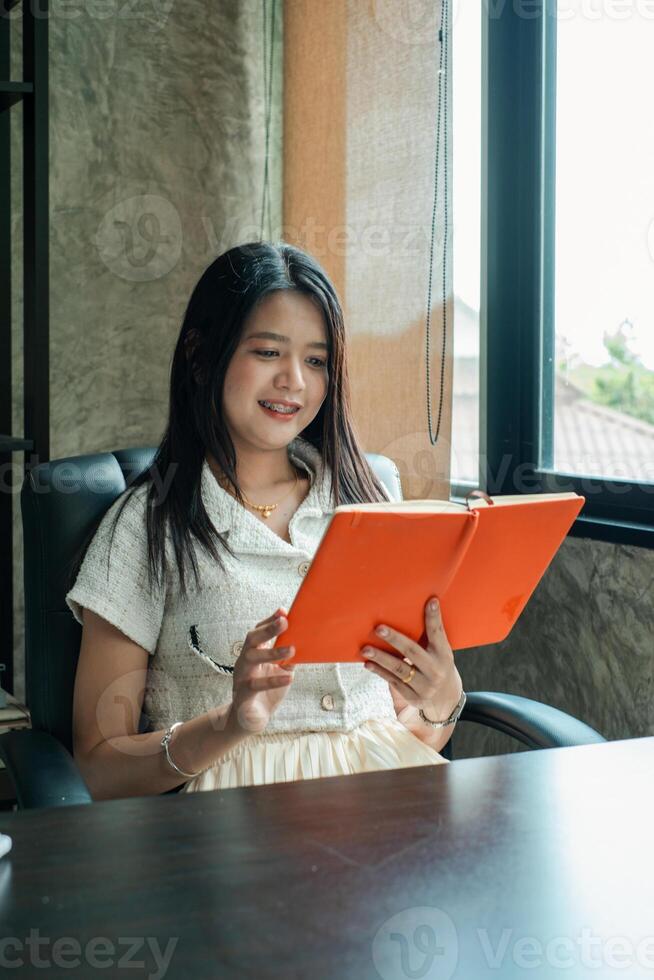 A woman enjoys reading an orange notebook while seated by a window in a contemporary office environment. photo