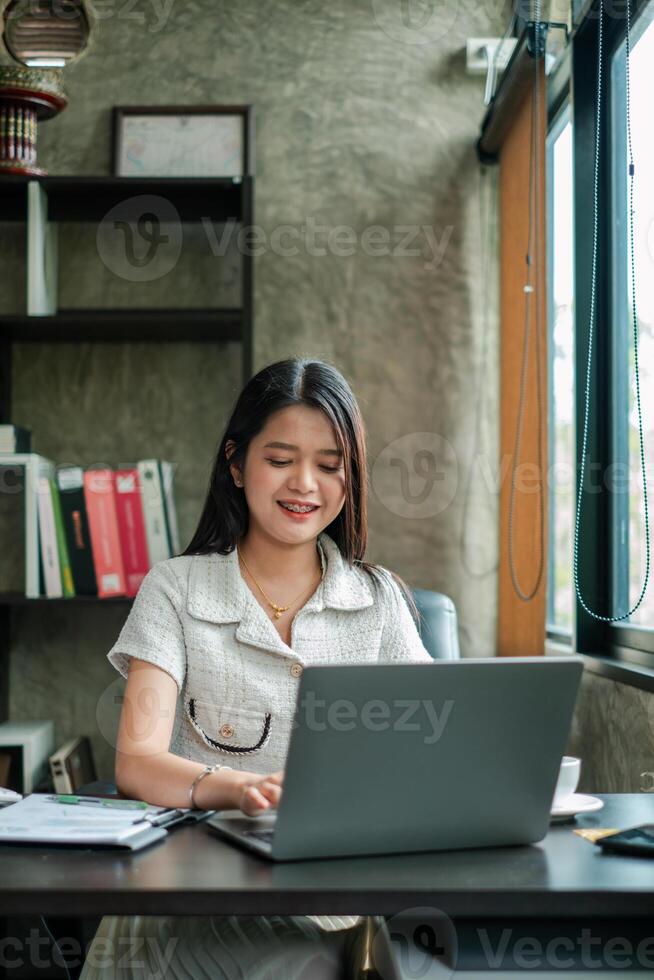 A young woman working on a laptop in a modern office setting, surrounded by documents and natural light from a large window. photo