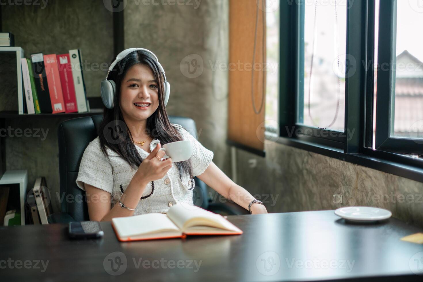 Smiling woman with headphones holding coffee cup, sitting at desk with open notebook in a bright office space. photo