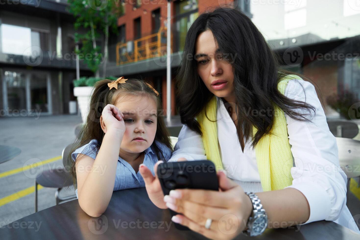 Mother and daughter enjoying time together while looking at a phone outside a modern building on a sunny day photo