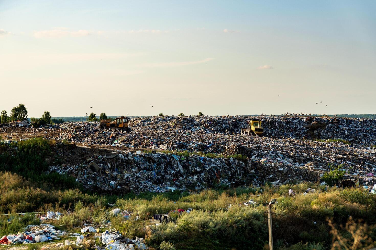 Heavy machinery works at a sprawling landfill filled with heaps of waste. The landscape is dotted with trees and a clear sky, indicating a warm, sunny day photo
