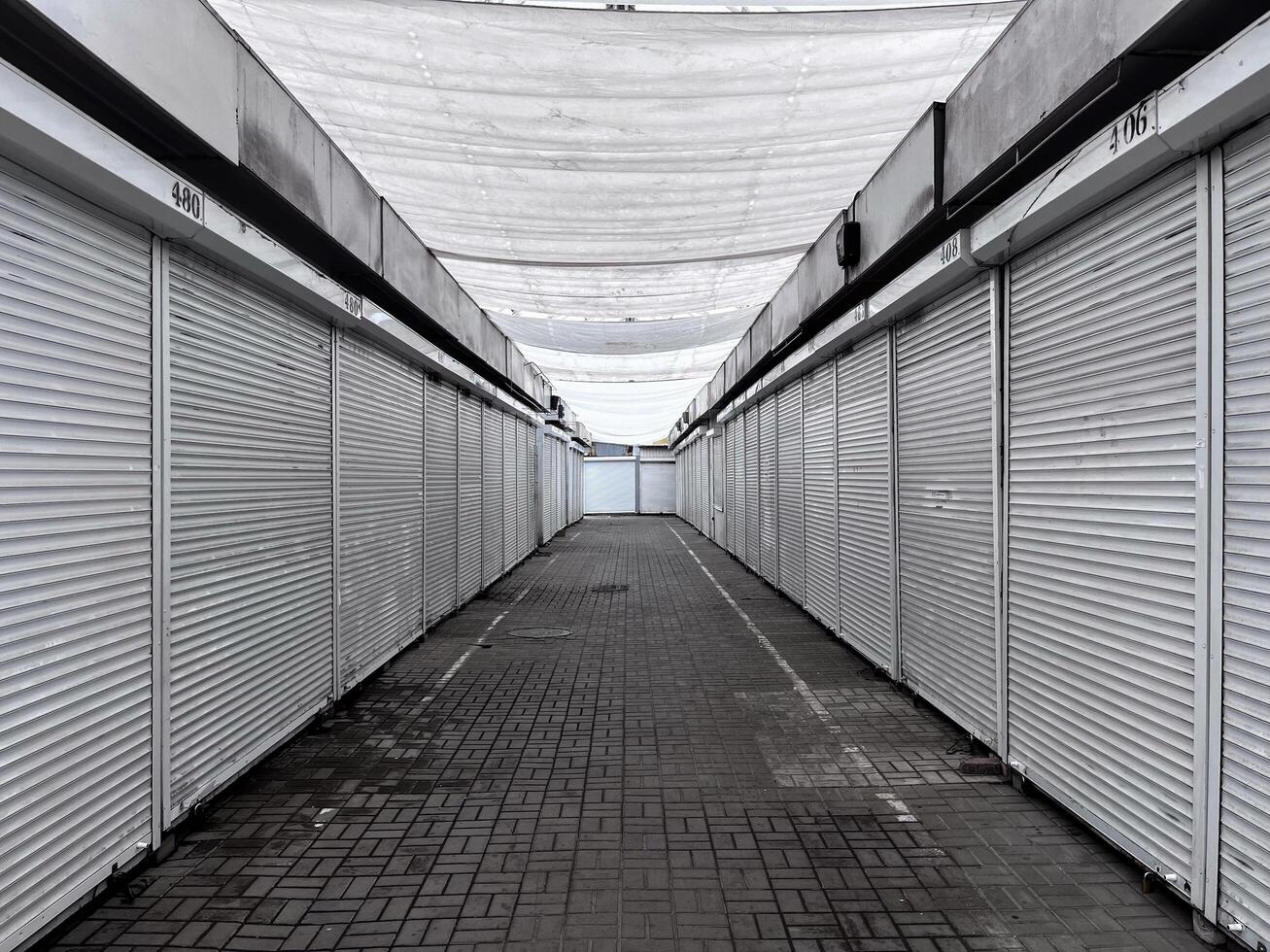 An empty marketplace features rows of closed stalls under a white canopy. The pavement is lined with tiles, and the atmosphere reflects a quiet afternoon in a commercial area photo