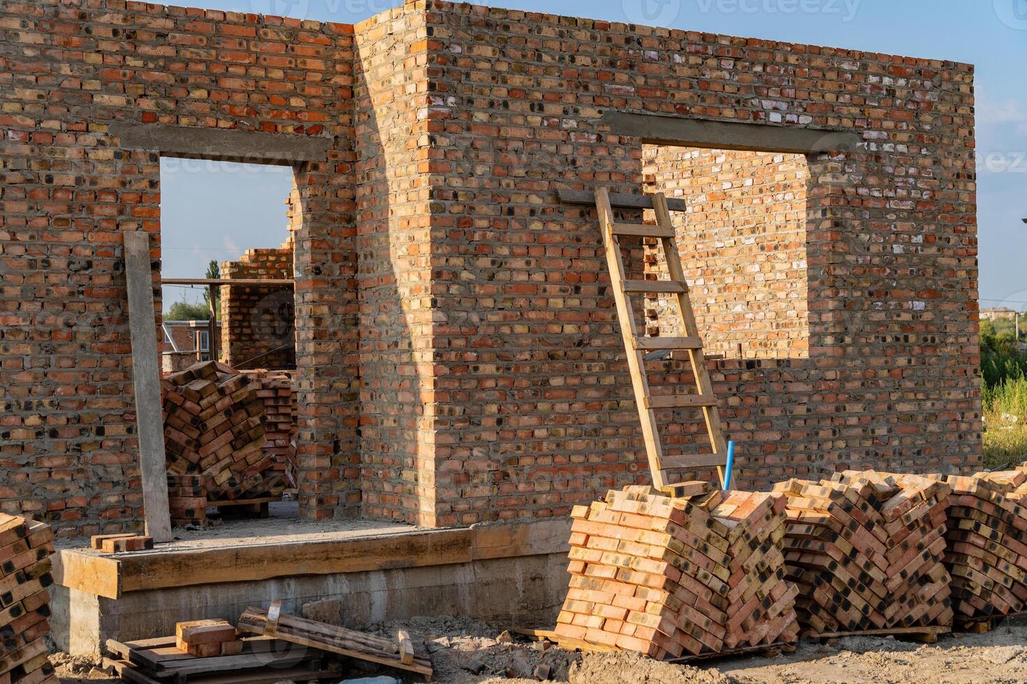 Construction workers are building a brick wall, which stands partially completed. A wooden ladder leans against the structure, and neatly stacked bricks nearby await use photo