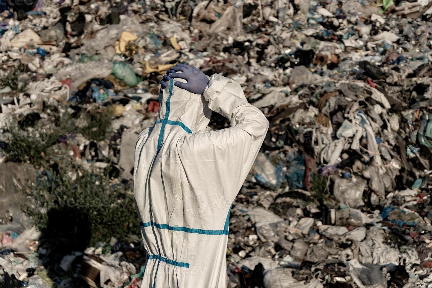 A worker wearing a protective suit examines a sprawling landfill overflowing with waste materials under bright daylight, highlighting the urgent issue of pollution and waste management photo